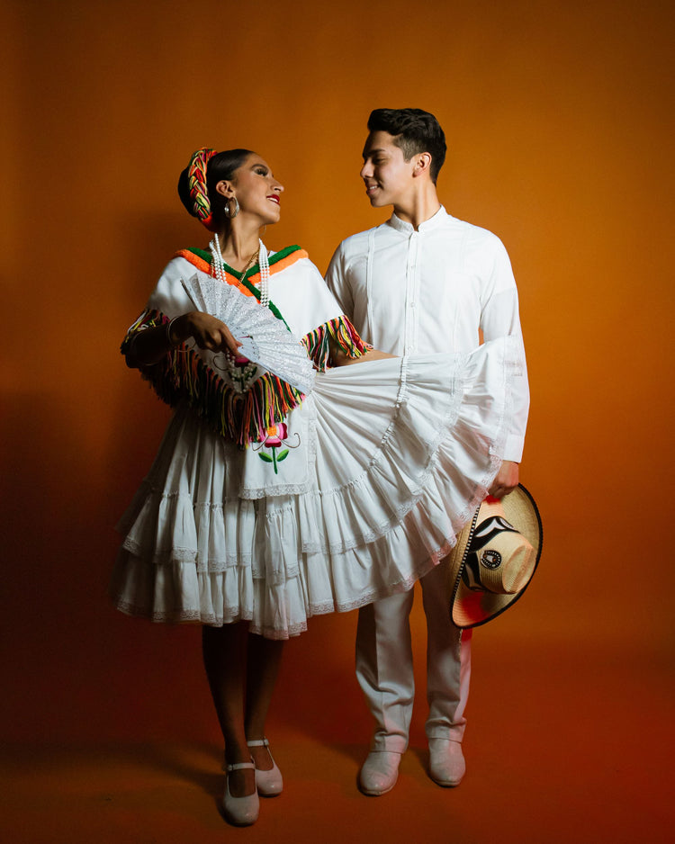 Folklórico couple in white Veracruz attire with dance shoes, against a warm orange backdrop